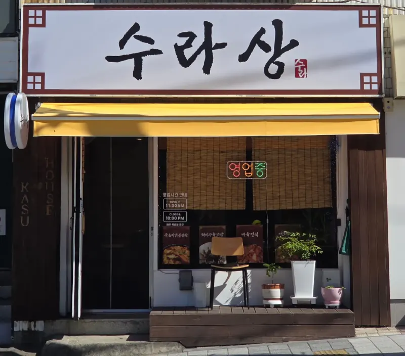 수라상 가게 외관. 흰 간판에 먹색 붓글씨 '수라상', 노란 차양, 창에 '영업중' 네온사인, 앞에 나무 데크